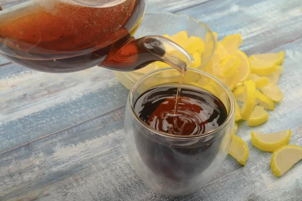 Le thé noir est versé d'une théière dans un verre, confiture tranches de citron sur un fond en bois. Gros plan . — Photo