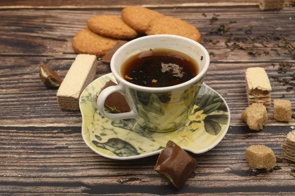 Una taza de té negro, hojas de té, trozos de azúcar morena, galletas de avena, gofres, trozos de chocolate sobre un fondo de madera. De cerca. . — Foto de Stock