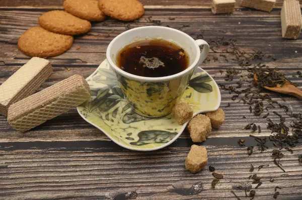 Una tazza di tè nero, foglie di tè, pezzi di zucchero di canna, biscotti di farina d'avena, cialde su uno sfondo di legno. Da vicino. . — Foto Stock