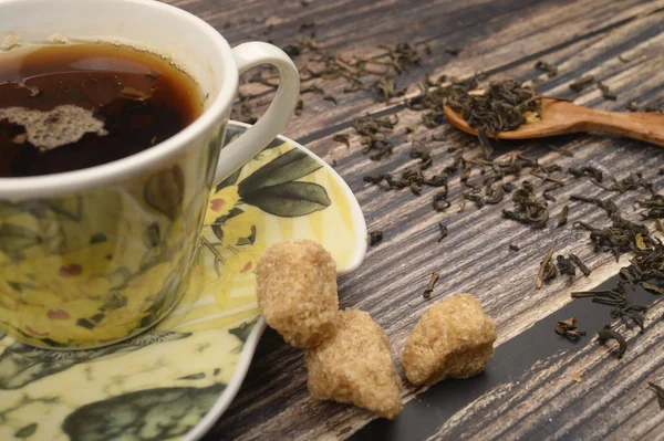 Чашка черного чая, чайные листья в деревянной ложке, кусочки коричневого сахара на деревянном фоне. Закрыть . — стоковое фото