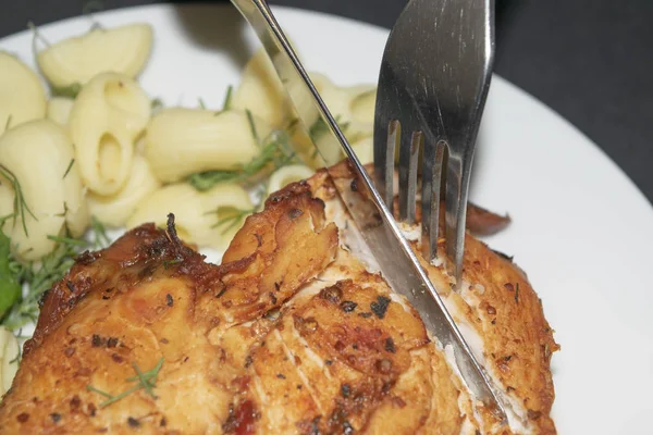 Kuřecí prsa smažená s kořením, vařené těstoviny s bylinkami, kovovou vidličkou a nožem, který krájí kus prsu na bílém talíři. — Stock fotografie