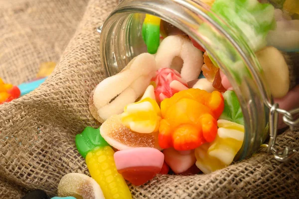 Multicolored shaped marmalade in a glass jar on a background of rough homespun fabric close-up. Delicious holiday treat. — Stock Photo, Image