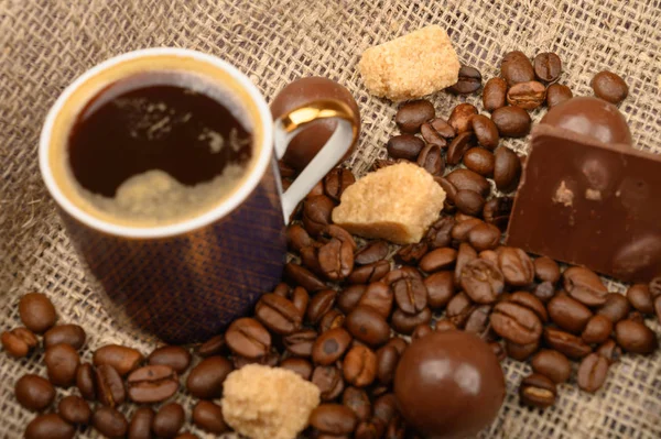 En kopp kaffe, kaffebönor, bitar av brunt socker och choklad på en bakgrund av grov hemspunnen tyg. Närbild. — Stockfoto