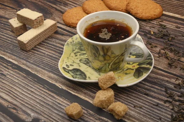 Eine Tasse schwarzen Tee, Teeblätter, braunen Zucker, Haferflockenkekse, Waffeln auf einem hölzernen Hintergrund. Nahaufnahme. — Stockfoto