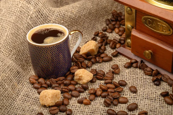 Чашка кофе, кофейные зерна, кусочки коричневого сахара и кофемолка на фоне грубой домашней ткани. Закрыть . — стоковое фото