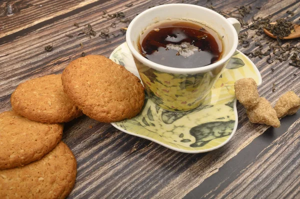 Una taza de té negro, hojas de té, trozos de azúcar morena, galletas de avena sobre un fondo de madera. De cerca. . — Foto de Stock