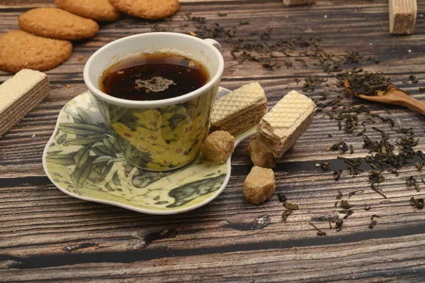 Una tazza di tè nero, foglie di tè, pezzi di zucchero di canna, biscotti di farina d'avena, cialde su uno sfondo di legno. Da vicino. . — Foto Stock