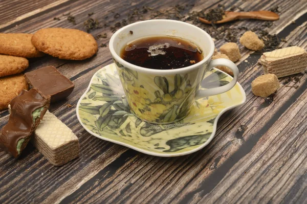 Una taza de té negro, hojas de té, trozos de azúcar morena, galletas de avena, gofres, trozos de chocolate sobre un fondo de madera. De cerca. . — Foto de Stock