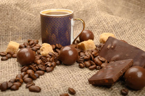 Een kopje koffie, koffiebonen, stukjes bruine suiker en chocolade op een achtergrond van ruwe homespun stof. Sluiten.. — Stockfoto