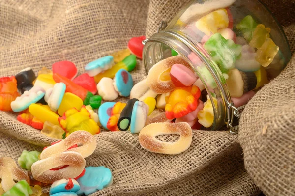 Multicolored shaped marmalade in a glass jar on a background of rough homespun fabric close-up. Delicious holiday treat. — Stock Photo, Image