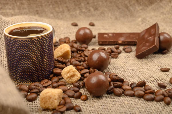 Eine Tasse Kaffee, Kaffeebohnen, brauner Zucker und Schokolade auf einem Hintergrund aus grobem selbstgesponnenem Stoff. Nahaufnahme. — Stockfoto