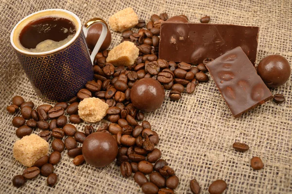 Eine Tasse Kaffee, Kaffeebohnen, brauner Zucker und Schokolade auf einem Hintergrund aus grobem selbstgesponnenem Stoff. Nahaufnahme. — Stockfoto