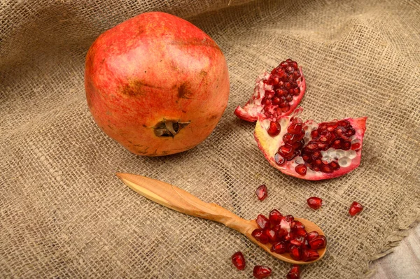 Ripe juicy pomegranate, pomegranate seeds in a wooden spoon and pieces of pomegranate on a background of rough homespun fabric. Close up. — Stock Photo, Image