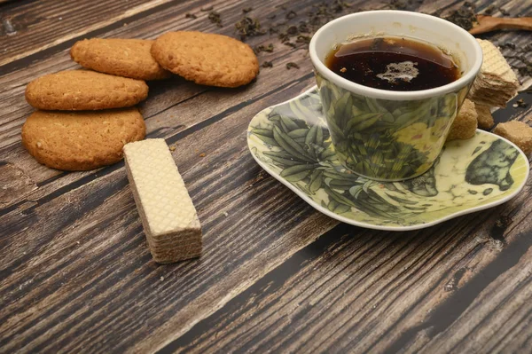 Eine Tasse schwarzen Tee, Teeblätter, braunen Zucker, Haferflockenkekse, Waffeln auf einem hölzernen Hintergrund. Nahaufnahme. — Stockfoto