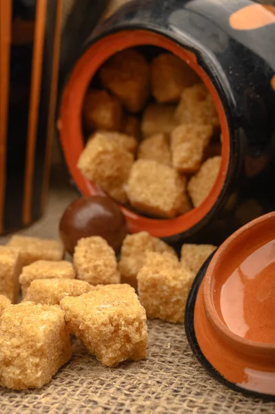 Pieces of brown sugar, a sugar bowl, and pottery against a background of coarse homespun cloth. Close up.