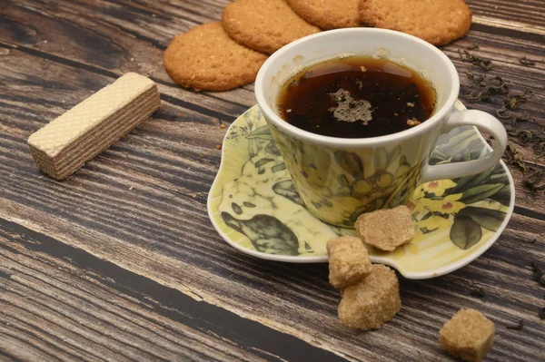 Eine Tasse schwarzen Tee, Teeblätter, braunen Zucker, Haferflockenkekse, Waffeln auf einem hölzernen Hintergrund. Nahaufnahme. — Stockfoto