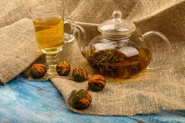 Цветочный чай, сваренный в стеклянном чайнике, стакан чая и шары цветочного чая на фоне грубой домашней ткани. Закрыть . — стоковое фото