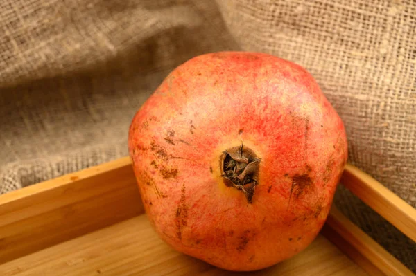 Ein reifer, saftiger Granatapfel auf einem hölzernen Tablett vor einem Hintergrund aus grobem, selbstgesponnenem Stoff. Nahaufnahme. — Stockfoto