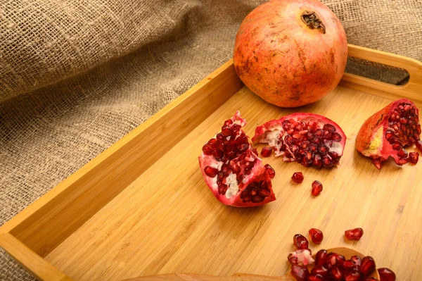 Reife saftige Granatäpfel, Granatapfelstücke, Granatapfelkerne auf einem Holzlöffel auf einem hölzernen Tablett auf einem Hintergrund aus grobem selbstgesponnenem Stoff. Nahaufnahme. — Stockfoto