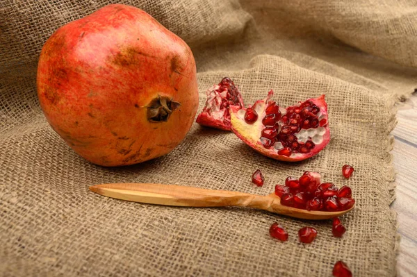 Ripe juicy pomegranate, pieces of pomegranate and pomegranate seeds on a wooden spoon on a background of rough homespun fabric. Close up. — Stock Photo, Image