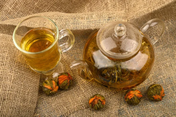 Blomma te bryggt i ett glas tekanna, ett glas te och bollar av blomma te på en bakgrund av grov hemspunnen tyg. Närbild. Stockfoto