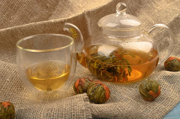 Flower tea brewed in a glass teapot, a glass of tea and balls of flower tea on a background of rough homespun fabric. Close up.