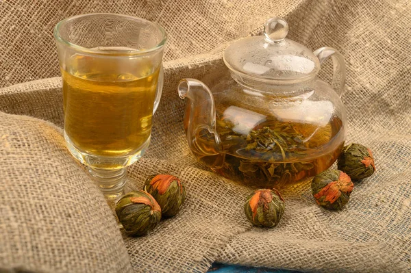 Flower tea brewed in a glass teapot, a glass of tea and balls of flower tea on a background of rough homespun fabric. Close up.