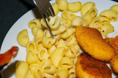 Çatallı birkaç haşlanmış makarna ve arka planda kızarmış tavuk nugget, makarna ve ketçap ile birlikte beyaz tabak. Kapat..