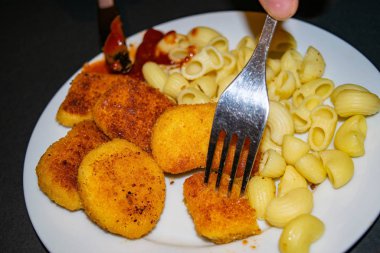 Çatallı bir parça nugget ve arka planda kızarmış tavuk nugget, makarna ve ketçap ile birlikte beyaz tabak. Kapat..