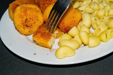 Çatallı bir parça nugget ve arka planda kızarmış tavuk nugget, makarna ve ketçap ile birlikte beyaz tabak. Kapat..