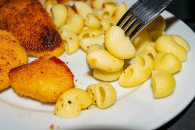 Çatallı birkaç haşlanmış makarna ve arka planda kızarmış tavuk nugget, makarna ve ketçap ile birlikte beyaz tabak. Kapat..