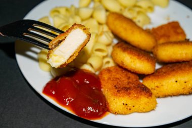Çatallı bir parça nugget ve arka planda kızarmış tavuk nugget, makarna ve ketçap ile birlikte beyaz tabak. Kapat..