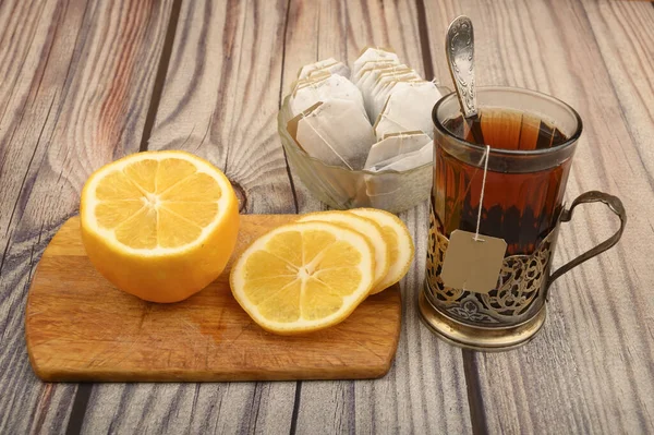 Thé dans un verre à facettes dans un porte-gobelet vintage, sachets de thé dans un vase en verre et citron tranché sur une planche sur un fond en bois. Gros plan . — Photo
