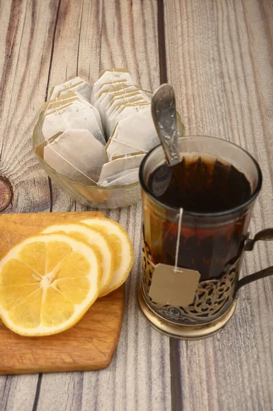 Thé dans un verre à facettes dans un porte-gobelet vintage, sachets de thé dans un vase en verre et citron tranché sur une planche sur un fond en bois. Gros plan . — Photo