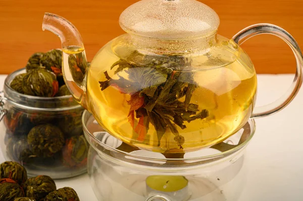Цветочный чай, сваренный в стеклянном чайнике, цветочные чайные шарики и банка чая на белом фоне. Закрыть . — стоковое фото