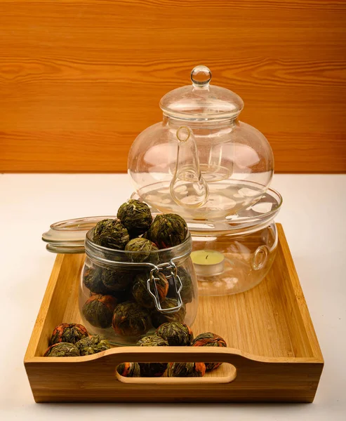 Flower tea balls in a glass jar and a glass teapot on a wooden tray on a white background. Close up. — Stock Photo, Image