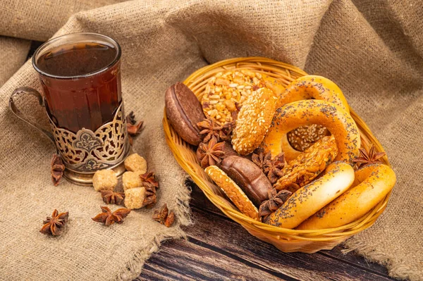 Cookies, bagels, chocolate cakes and star anise in a wicker basket and a glass of tea in a vintage Cup holder on a background of rough homespun fabric. Close up