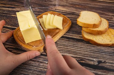 Birisi tereyağını, ahşap bir tereyağından bir parça bıçakla buğday ekmeğinden taze tost ve ahşap bir zeminde peynir dilimleriyle keser. Kapat.