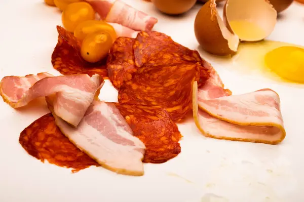 Ovo de galinha quebrado, fatias de linguiça e bacon e tomates em um fundo branco. Fechar . — Fotografia de Stock