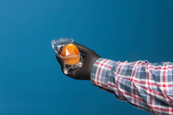 Industrial cupcake in the hand disinfected and protected with a black glove, on a blue background. Industrial pastries