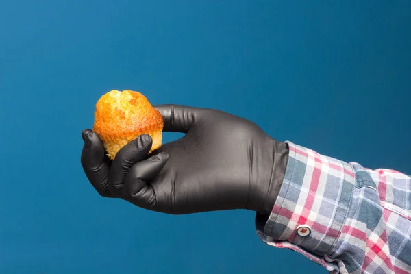Industrial cupcake in the hand disinfected and protected with a black glove, on a blue background. Industrial pastries