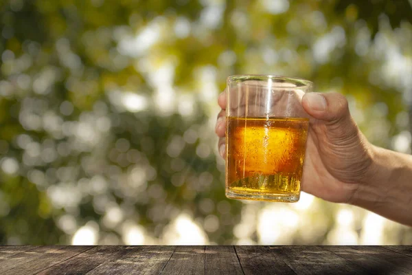 Hands are lifting a glass of beer from a wooden table and natural bokeh background.