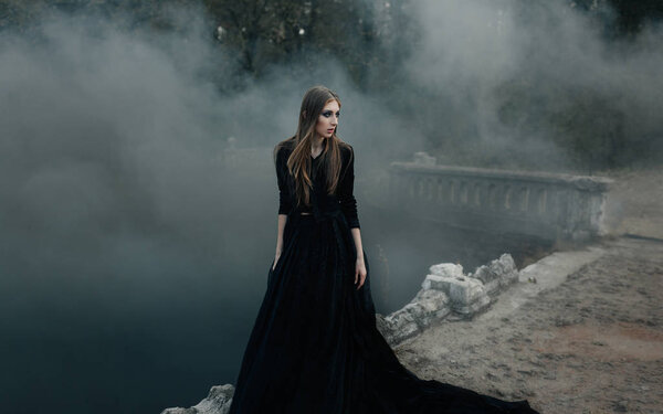 Young attractive Witch in black dress walking on the old bridge in heavy black smoke. Dark magic. Horror fairy tale.