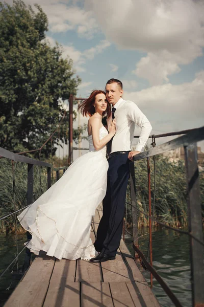 Casal de casamento andando na ponte perto do lago ao pôr do sol no dia do casamento. Noiva e noivo apaixonados — Fotografia de Stock