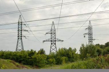 Güneşli bir günde düz ve yeşil bir tarım arazisinde yüksek voltaj hatları ve elektrik direkleri.