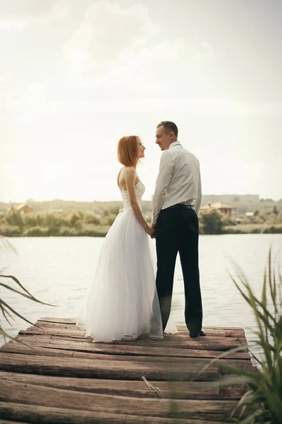 Pareja de boda caminando en el puente cerca del lago en la puesta del sol en el día de la boda. Novia y novio enamorados — Foto de Stock