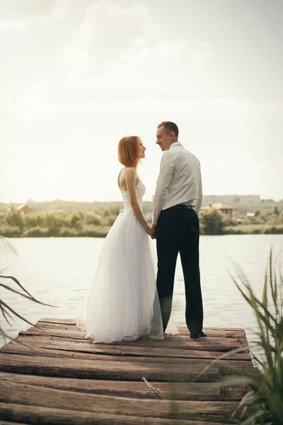 Casal de casamento andando na ponte perto do lago ao pôr do sol no dia do casamento. Noiva e noivo apaixonados — Fotografia de Stock