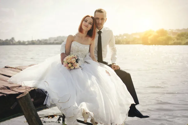 Casamento casal sentado na ponte perto do lago no pôr do sol no dia do casamento. Noiva e noivo apaixonados — Fotografia de Stock