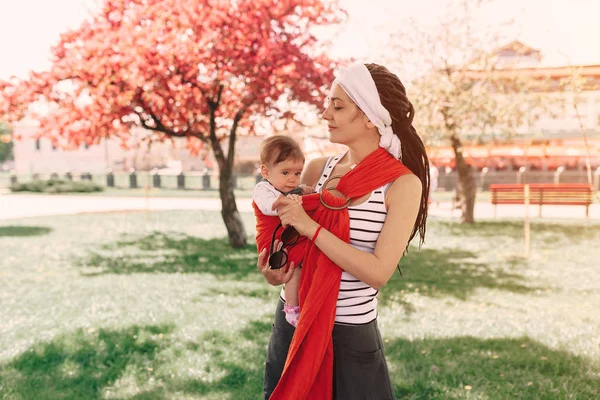 Мать носит младенца в рогатке в парке. Весной. Понятие естественного воспитания — стоковое фото