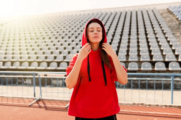 Hermosa dama corredora en ropa de abrigo en el estadio mirando a un lado. Usar capucha roja . — Foto de Stock